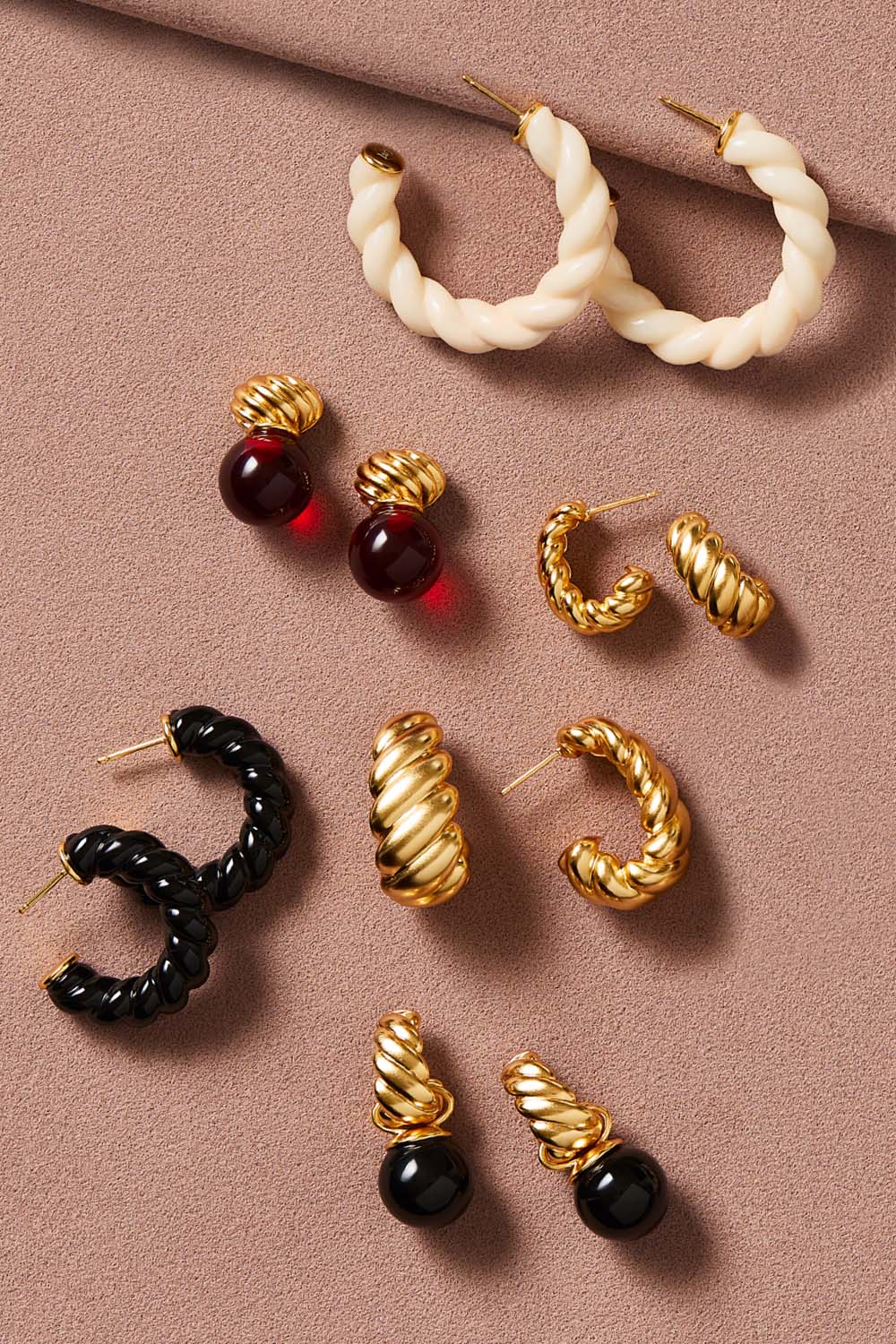 A styled flat-lay of multiple pairs of statement hoop and curb earrings arranged on a dusty pink background. The earrings include cream rope-textured hoops, black rope-textured hoops, gold curb hoops with twisted details, and pairs with dark red and black glossy bead accents.