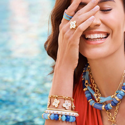 A close-up lifestyle shot of a woman smiling near a swimming pool, showcasing a stack of blue and gold bracelets on her wrist, a matching blue beaded necklace, and a gold flower ring on her finger.