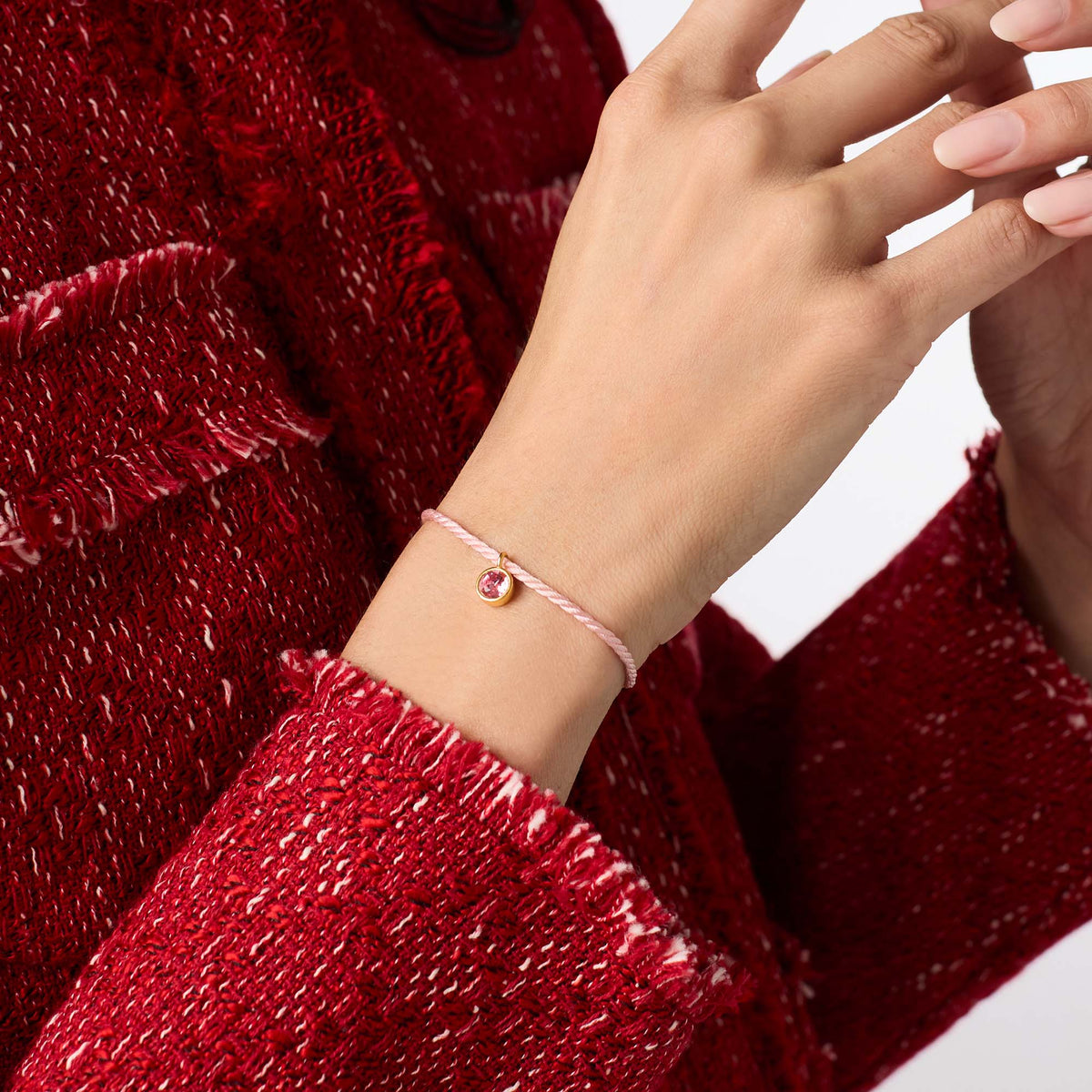 carousel, A close-up of a person's wrist wearing a delicate pink cord bracelet with a small gold bezel-set pink gemstone, styled with a textured red tweed jacket.
