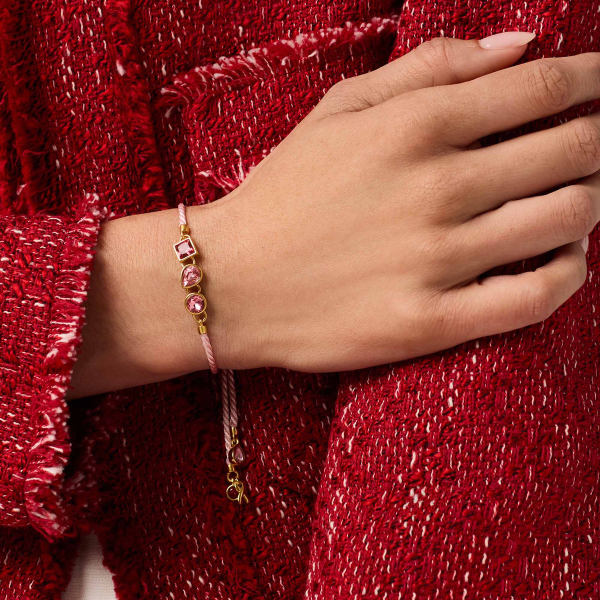 carousel, A close-up of a person's hand and wrist wearing a delicate pink cord bracelet with gold-toned hardware and pink gemstone beads, set against a textured red tweed fabric background.